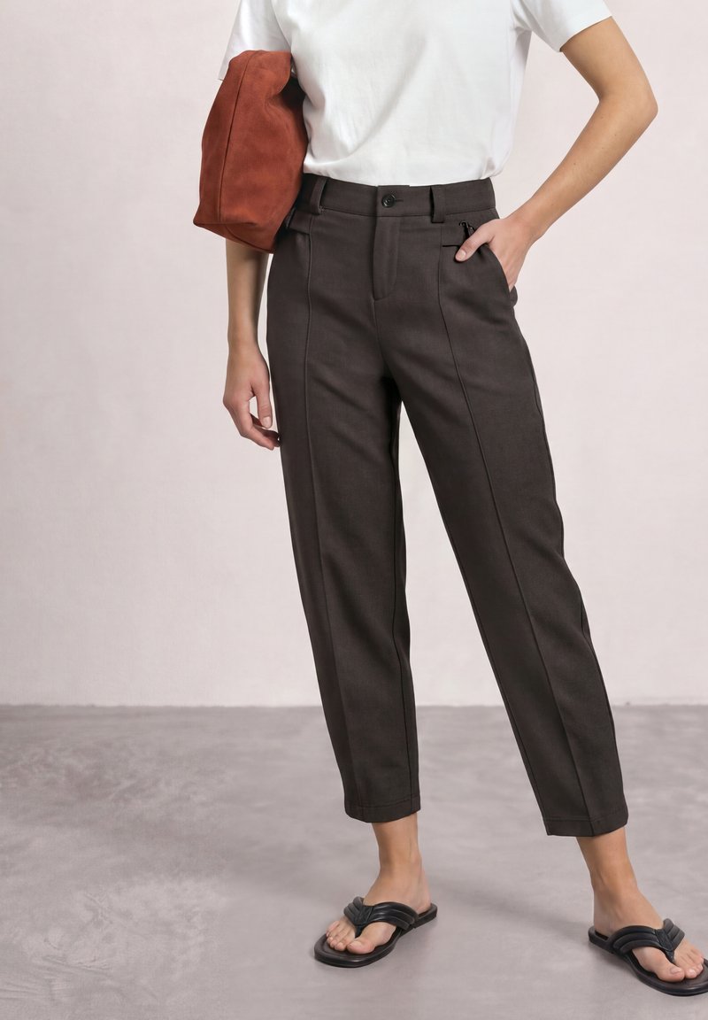 Person wearing high-waist dark brown tapered pants, white t-shirt, black slip-on sandals, and holding a rust-colored soft bag under one arm.