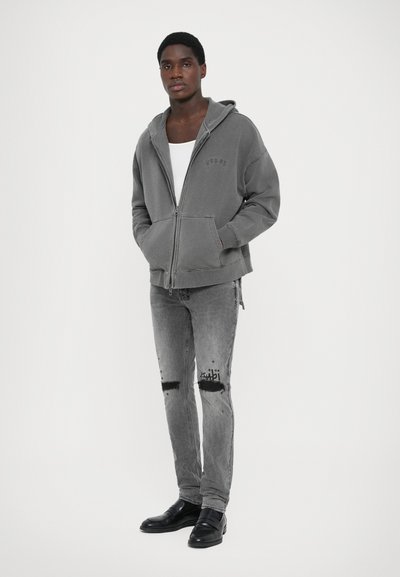 Grå zip-up hoodie med huva, featuring framfickor och en subtil logotyp. Bärs med slitna grå jeans och svarta skor.