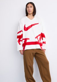 Unge person med langt hår iført en overdimensioneret hvid sweater med et stort rødt Nike-logo og brune løse bukser, stående mod en hvid væg.