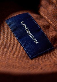Bruine gestructureerde stof met een donkerblauw label met daarop "LINDBERGH" in het wit, met gestikte randen. Close-up opname legt de details van het materiaal bloot.