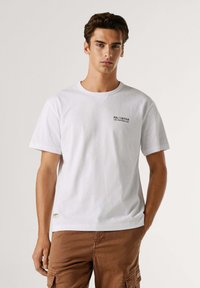 Wit katoenen T-shirt met korte mouwen, ronde hals en een klein zwart bedrukt ontwerp op de linkerborst. Minimalistische stijl.