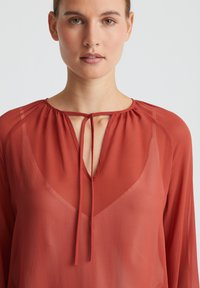 Rote durchsichtige Bluse mit rundem Ausschnitt, die eine Bindeschließe und lange Ärmel hat. Leichter, fließender Stoff mit einem subtilen Glanz.