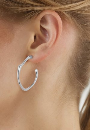 Orecchio con un orecchino a cerchio aperto di forma irregolare in argento, indossato da una persona con pelle chiara e capelli biondi raccolti.