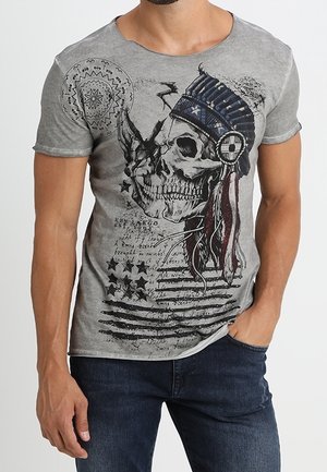 T-shirt gris à manches courtes avec un graphique de tête de mort portant une coiffe en plumes bleues, accompagné d'un texte et d'un design de drapeau américain usé.