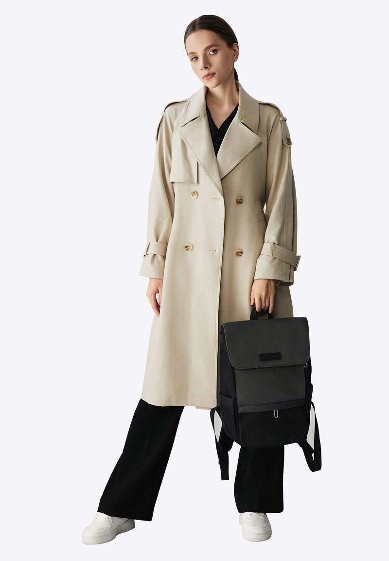 Beige dubbelknäppt trenchcoat med guldknappar, svart topp, vida svarta byxor, vita sneakers och en svart ryggsäck med lock.