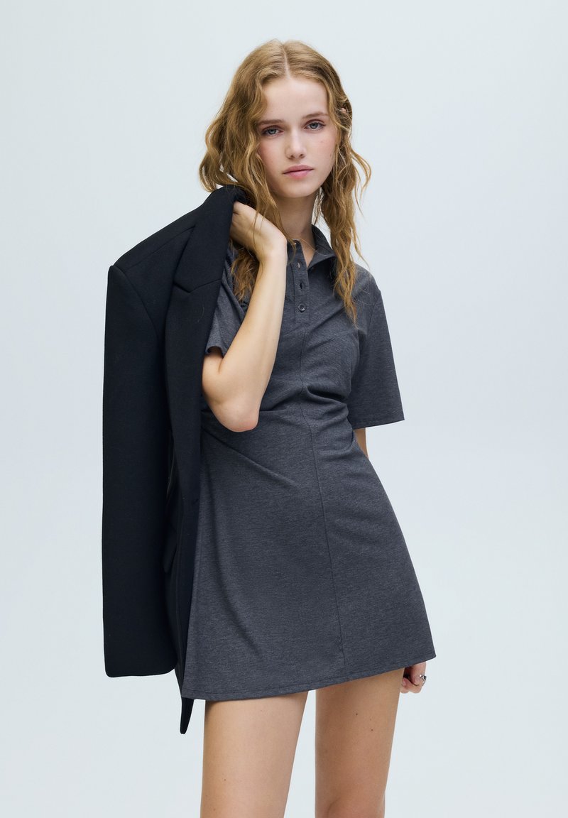 Szürke, testhez simuló szabású pólóruha rövid ujjakkal és gombos elülső résszel. A modell fekete blézert tart a vállán egy semleges háttér előtt.