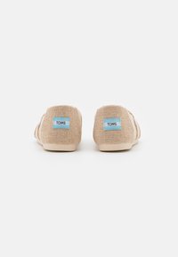 Chaussures à enfiler en tissu tan avec des semelles beige clair, présentant une petite étiquette TOMS bleue à l'arrière. Texture lisse, forme arrondie.