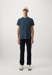 Blue short-sleeve t-shirt, dark jeans, and white sneakers with tan accents. Model wears a light beige cap, standing against a plain background.