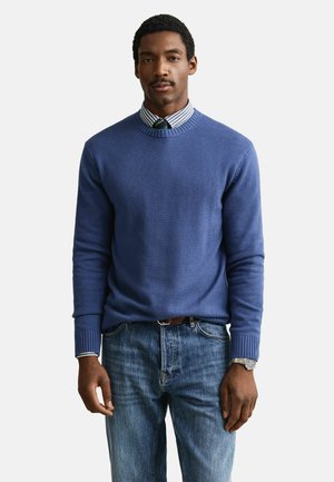 Man draagt een blauwe trui over een gestreept overhemd met stropdas, blauwe spijkerbroek en polshorloge, staand tegen een effen witte achtergrond.