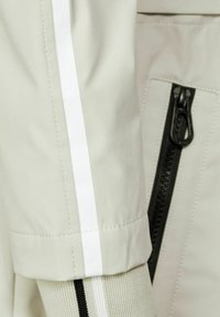 Světle béžová bunda s bílým pruhem, která má černý zip na boku a texturované pletené manžety v kontrastní světle barvě.