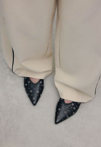 Black leather pointed-toe shoes with silver grommet accents, worn with cream-colored wide-leg trousers. Smooth texture on the shoe surface.