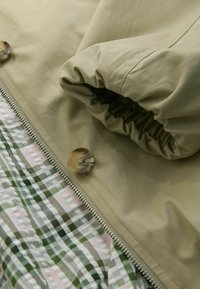 Geacă verde măsliniu cu un finisaj texturat, cu mânecă strânsă și un interior cu model în carouri în nuanțe de verde și roz. Detaliu cu nasturi vizibil.