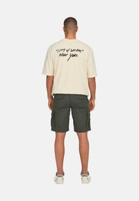 T-shirt oversized beige con testo nero "CITY OF DREAMS" e "New York" sul retro. Indossato con pantaloni cargo verde scuro e sneakers bianche.