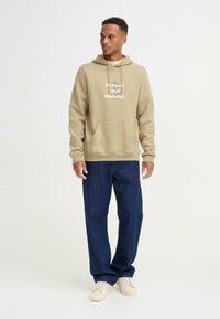 Sudadera beige con un bolsillo frontal y el gráfico "EXPAND YOUR MINDSET", combinada con jeans de mezclilla azul oscuro y zapatillas de color claro.