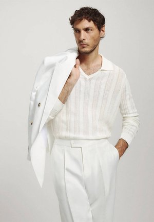 Homme aux cheveux courts et bouclés portant un pull en maille blanc et un pantalon blanc, tenant une veste blanche sur son épaule, regardant sur le côté.