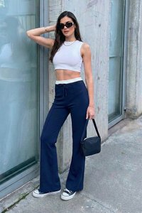 Jeune femme en crop top blanc, pantalon flare bleu marine, lunettes de soleil noires, baskets, tenant un petit sac à main noir, debout près d'un mur en verre et en béton.