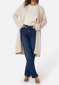 Beżowy kardigan otwarty z przodu, z fakturowanej tkaniny, z długimi rękawami nałożony na kremowy sweter. Zestawiony z ciemnoniebieskimi dżinsami o wysokim stanie i przezroczystymi sandałami.