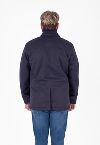 Giacca impermeabile blu navy con colletto alto, tessuto testurizzato e polsini con bottoni. Abbinata a jeans di denim blu. Vista posteriore.