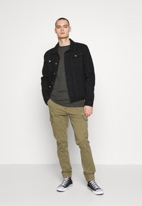 Svart denimjacka, grönrandig långärmad skjorta, olivgröna cargobyxor och svarta sneakers. Modellen står med en hand i fickan.