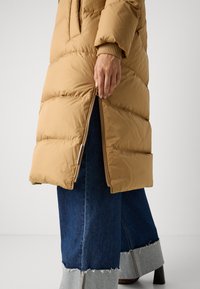 Bronzová pufovaná bunda s prešívaným dizajnom, zipsom a rebrovanými manžetami, v kombinácii s rozšírenými džínsami z denimu s rozstrapkaným lemom.