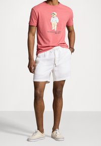 Rotes Baumwoll-T-Shirt mit einem Grafikdruck eines Bären in einem weißen Outfit, kombiniert mit weißen Leinen-Shorts und beigen Sneakers. Einfaches Design, lässiger Look.