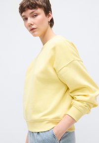 Κίτρινο φούτερ με άνετη εφαρμογή, ριμπ μανσέτες και στρίφωμα. Κατασκευασμένο από απαλό ύφασμα, με χαλαρή γραμμή στους ώμους και στρογγυλή λαιμόκοψη.