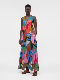 Femme portant une robe longue sans manches avec des motifs floraux et feuillus abstraits et colorés en rouge, bleu, rose, vert et marron, associée à des sandales marron.
