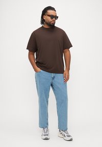 Καφέ κοντομάνικη μπλούζα συνδυασμένη με ανοιχτό μπλε τζιν παντελόνι με ίσια μπατζάκια και γκρι αθλητικά παπούτσια. Αξεσουάρ περιλαμβάνουν σκούρα γυαλιά ηλίου.