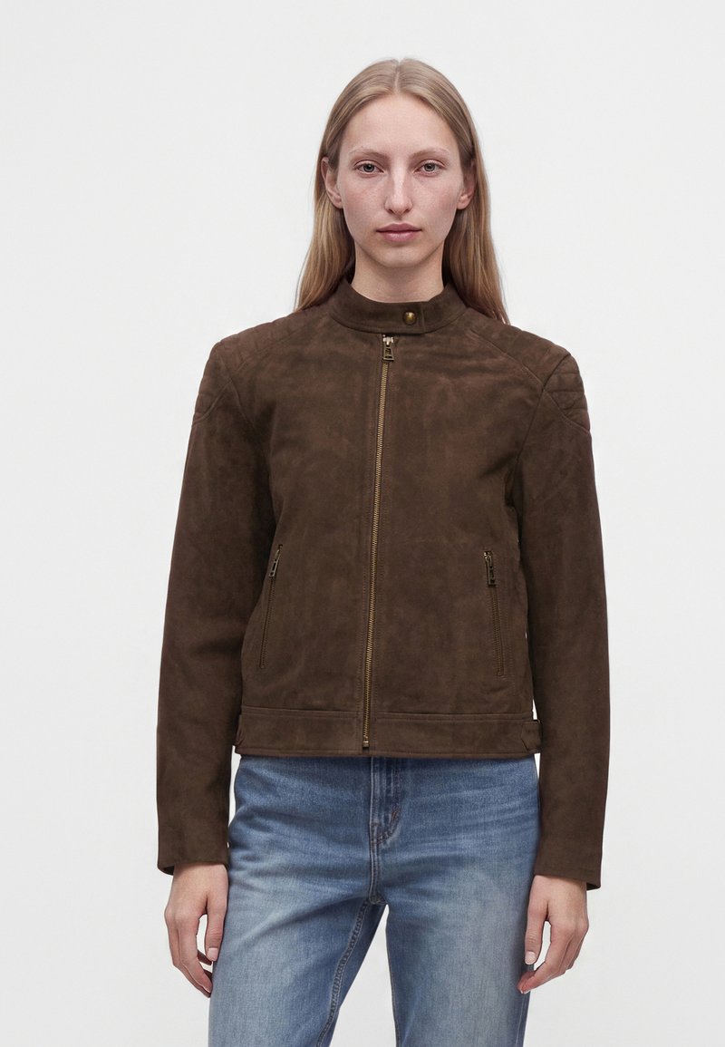 Femme aux cheveux blonds raides portant une veste zippée en daim marron et un jean bleu, debout devant un fond clair uni.