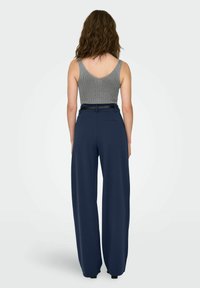 Naine, kellel on lainelised juuksed, kannab halli ilma varrukateta topi ja tumesiniseid avarate pükste, millel on must vöö, seistes seljaga tühja tausta poole.