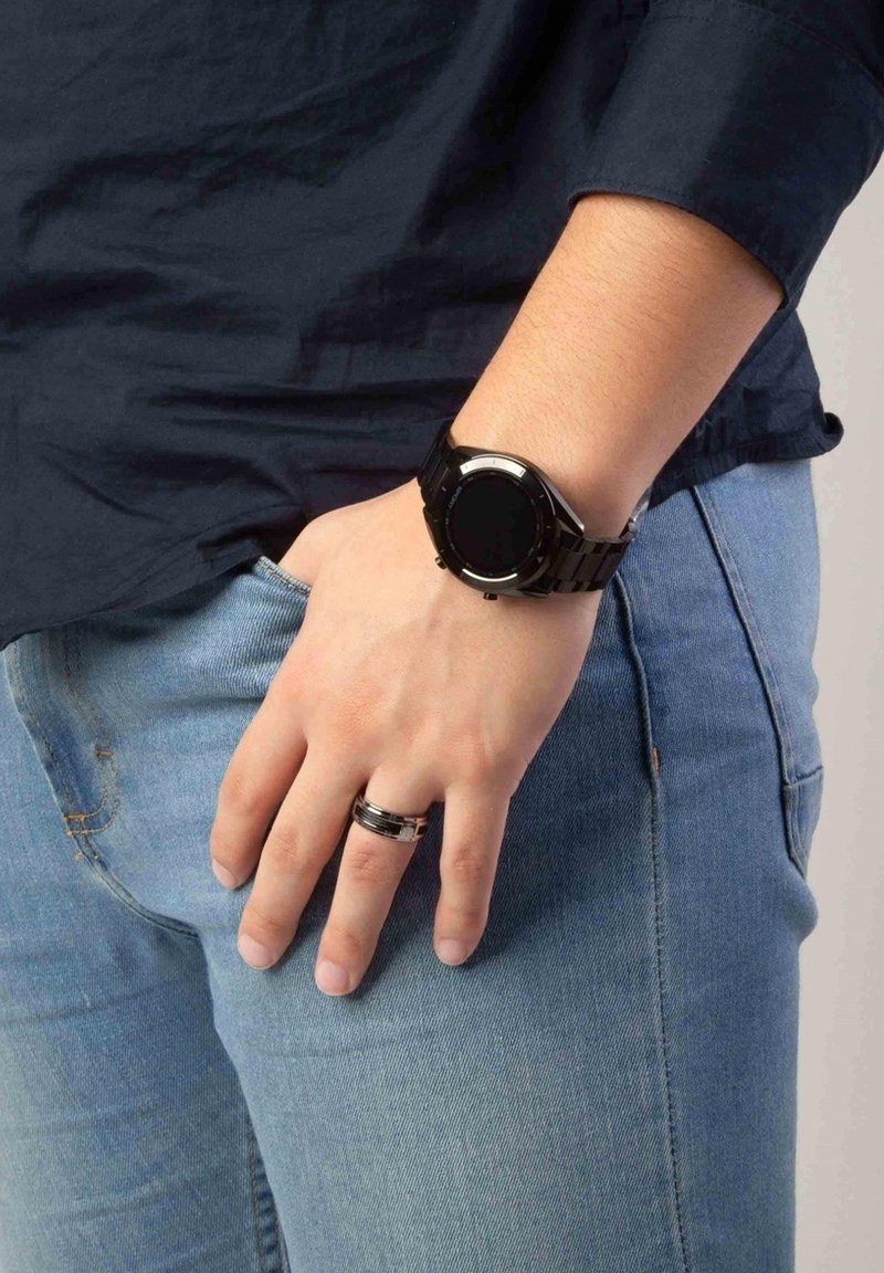 Black digital watch with a round face and textured strap, paired with a silver and black ring, worn on a relaxed, denim-clad hand.