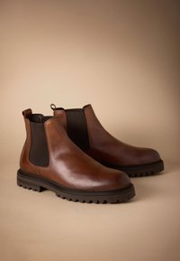 Hnedé kožené Chelsea topánky s elastickými bočnými panelmi a pútkami, vybavené robustnou tmavou gumovou podrážkou a hladkým, lesklým povrchom.