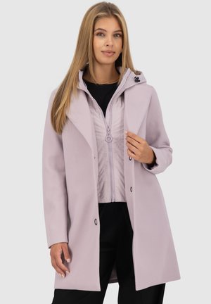 Une jeune femme aux cheveux blonds raides porte un manteau violet clair par-dessus une veste zippée et un pantalon noir, posant devant un fond uni.