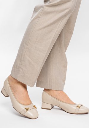 Beige flache Lederschuhe mit goldener Schnallendetailierung, getragen zu hellbeigen maßgeschneiderten Hosen auf weißem Hintergrund.