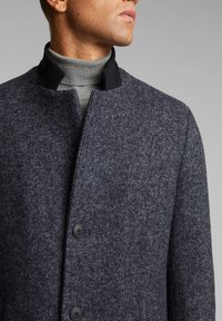 Uomo che indossa un cappotto in spina di pesce grigio scuro con colletto nero sopra un dolcevita grigio chiaro, su uno sfondo grigio uniforme.