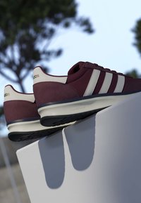 Baskets Adidas bordeaux et blanches avec semelles Cloudfoam reposant sur un piédestal blanc en extérieur avec des arbres flous en arrière-plan.