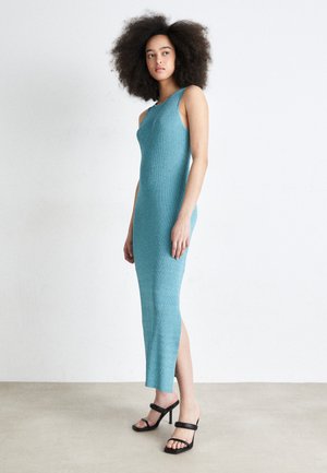 Een getailleerde, mouwloze jurk van een zachte, gestructureerde turquoise brei stof, met een zijsplit en gedragen met zwarte sandalen met bandjes.