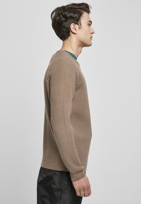 Maglione a coste marrone con maniche lunghe e scollo a girocollo, aderente al corpo. Abbinato a pantaloni scuri, profilo laterale mostrato.