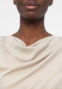 Κοντινό πλάνο του κάτω μέρους του προσώπου και του λαιμού μιας γυναίκας που φοράει μικρά χρυσά σκουλαρίκια και ένα ανοιχτό μπεζ τοπ από υφασμάτινη ύφανση.