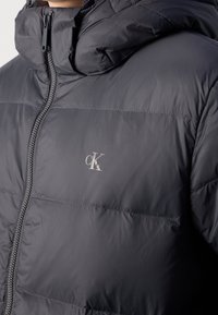 Veste matelassée noire avec col montant, fermeture éclair et détail logo. Texture lisse avec lignes de coutures et design épuré et moderne.