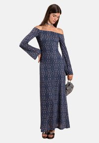 Langes, schulterfreies Kleid in Marineblau mit Blumenmuster, mit weit ausgestellten Ärmeln und einer figurbetonten Silhouette. Kombiniert mit einer kleinen grauen Handtasche und schwarzen Schuhen.