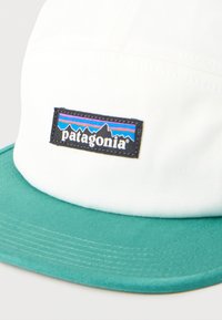 Λευκό και τιρκουάζ καπέλο Patagonia με ορθογώνια ετικέτα λογότυπου που απεικονίζει βουνά και ρίγες σε μπλε και μοβ.