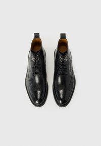 Stivali alla caviglia in pelle nera con design brogue, punte a goccia, perforazioni decorative, lacci neri e suola testurizzata.