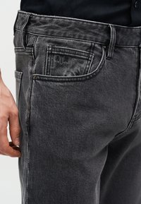 Κοντινή λήψη ενός ατόμου που φοράει τζιν σκούρου γκρι με φανερή μπροστινή τσέπη και ένα χέρι που ακουμπά κοντά στο μηρό.