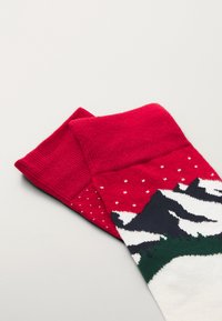 Sarkanas adītas zeķes ar ziemas kalnu ainavu baltā un zaļā krāsā, ar maziem baltajiem punktiem, kas atgādina sniega pārklājumu.