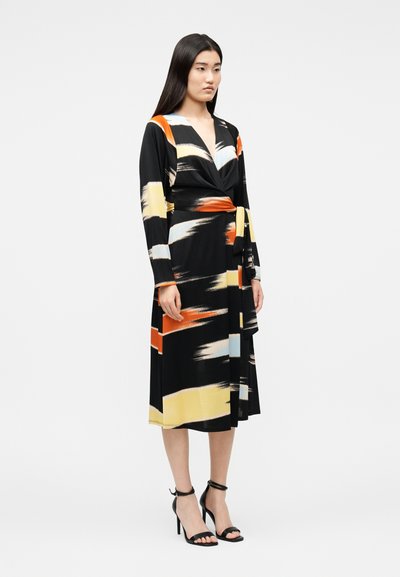 Γυναίκα με μαύρο φόρεμα τύπου wrap με μοτίβα σε πορτοκαλί, κίτρινο και ανοιχτό μπλε χρώμα, φοράει μαύρα σανδάλια με ψηλά τακούνια, στέκεται μπροστά σε απλό φόντο.