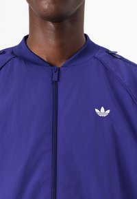 Lila lättvikts zip-up jacka med ribbad krage och vitt logo på bröstet. Har tryckknappar på axeln. Slät textur.