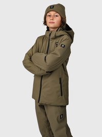 Ragazzo teenager con le braccia incrociate, indossa una giacca invernale verde oliva, pantaloni abbinati e un berretto su uno sfondo grigio uniforme.