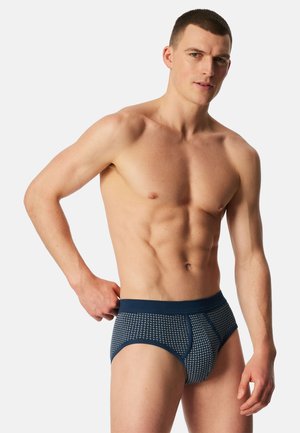 Man zonder shirt met kort haar, gekleed in blauwe, met patronen bedrukte slip, staand tegen een effen lichte achtergrond, hand rustend op de taille.