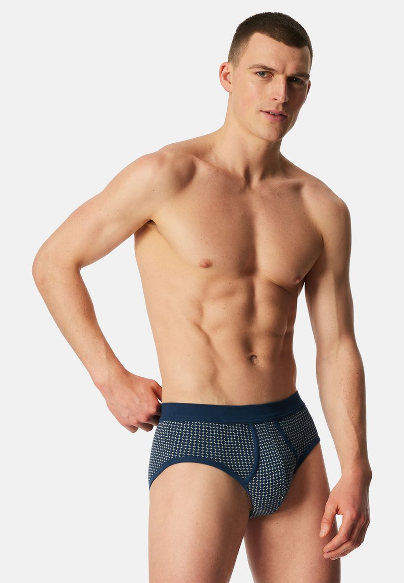 Paita pois päältä oleva lyhyttukkainen mies, jolla siniset kuvioidut alushousut, seisoo vaaleaa taustaa vasten, käsi lepää vyötäröllä.